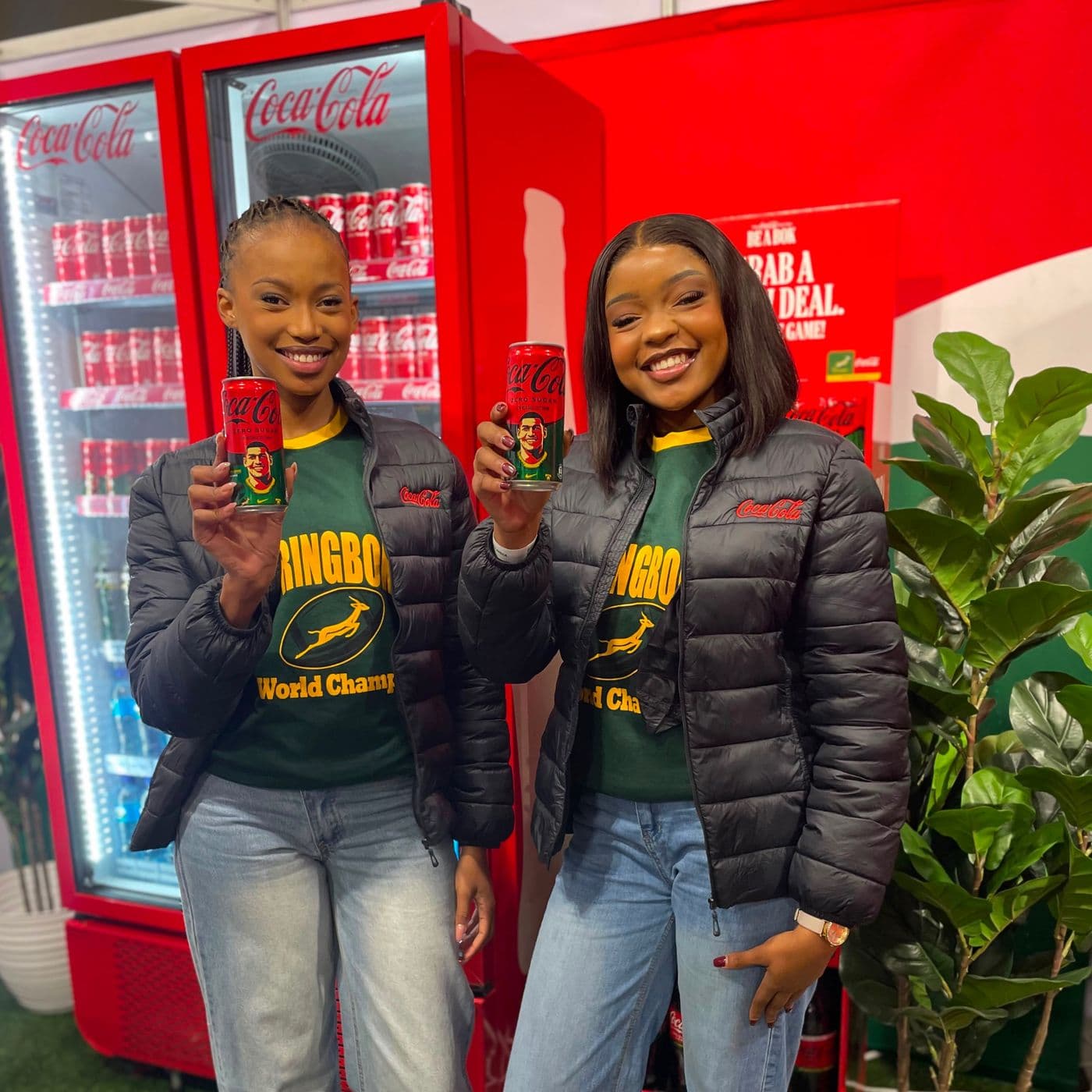 Two brand ambassadors in Springbok jerseys holding Coca-Cola cans beside branded fridges