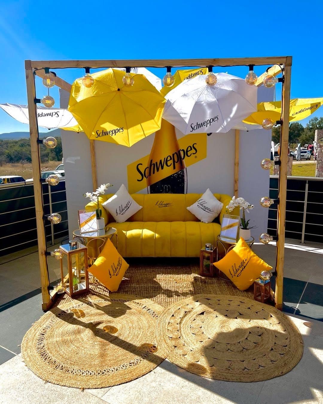 Schweppes lounge with yellow and white umbrellas overhead, a yellow sofa and a woven rug